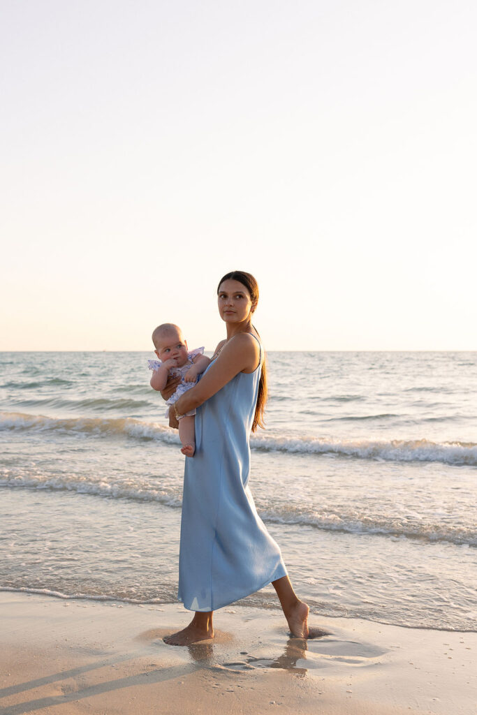 Mom walking on beach with feet in water while holding baby showcasing family beach photography tips.